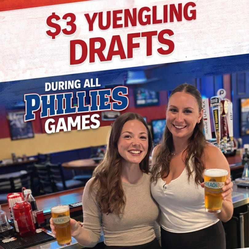 Two smiling women holding beers at a bar, with a promotional sign for $3 Yuengling drafts during Phillies games.