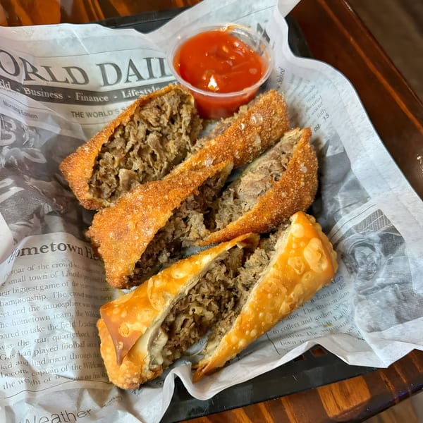 Four crispy egg rolls filled with shredded meat, served with a side of red dipping sauce on newspaper-lined tray.